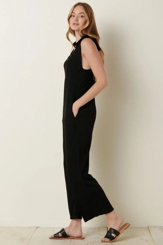 Woman in sleeveless black jumpsuit with side pockets and black sandals, standing indoors