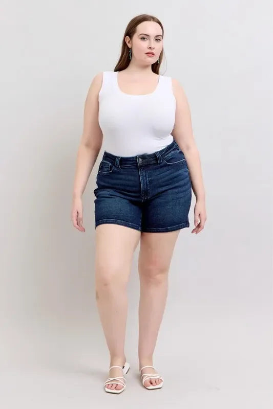 Woman in white tank top and dark denim shorts, standing against a plain background.
