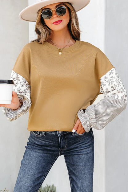 Woman in tan oversized sweatshirt with floral and striped sleeves, jeans, and sun hat