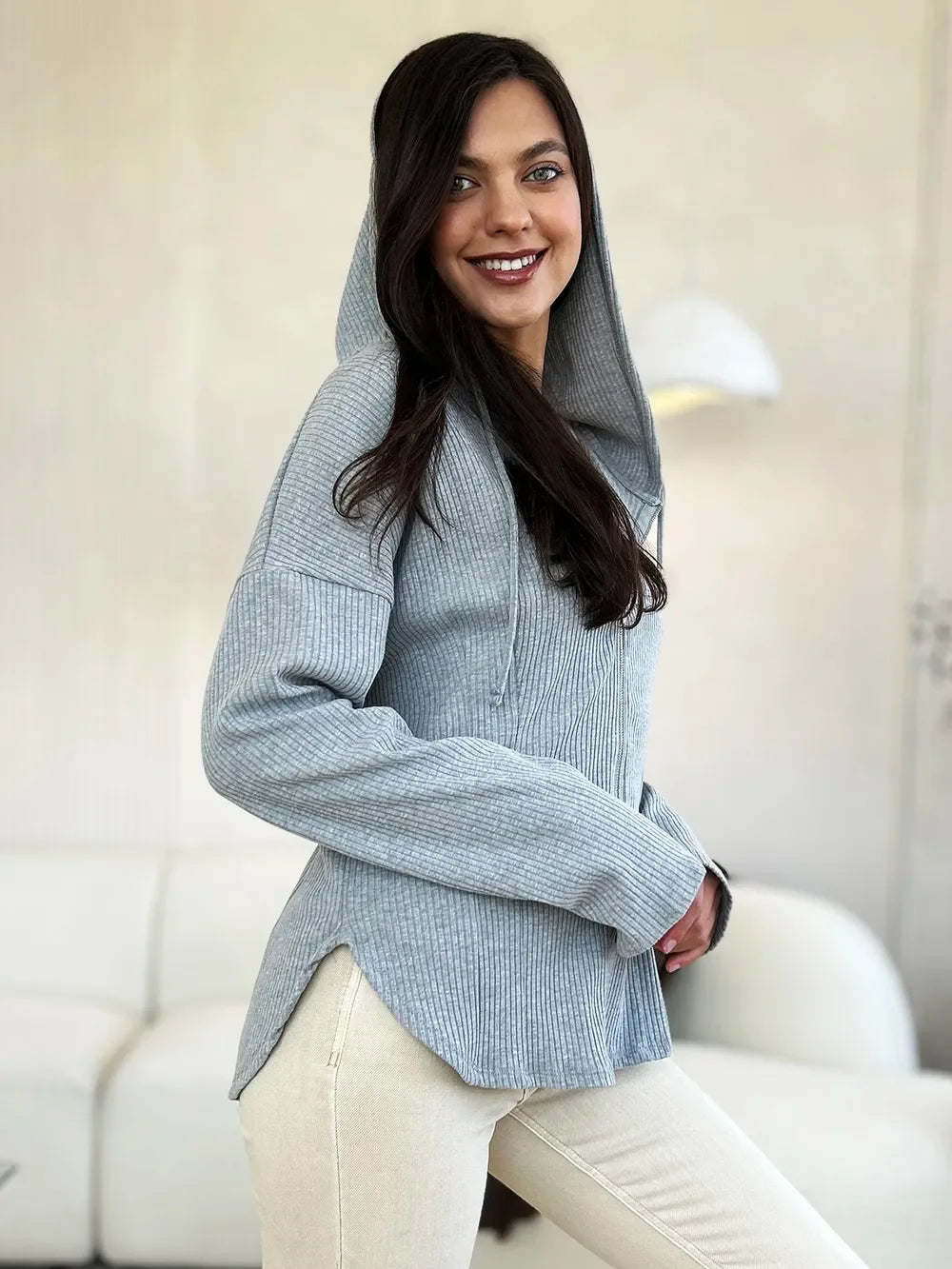 Woman wearing a light gray ribbed knit hoodie and cream pants in a modern living room