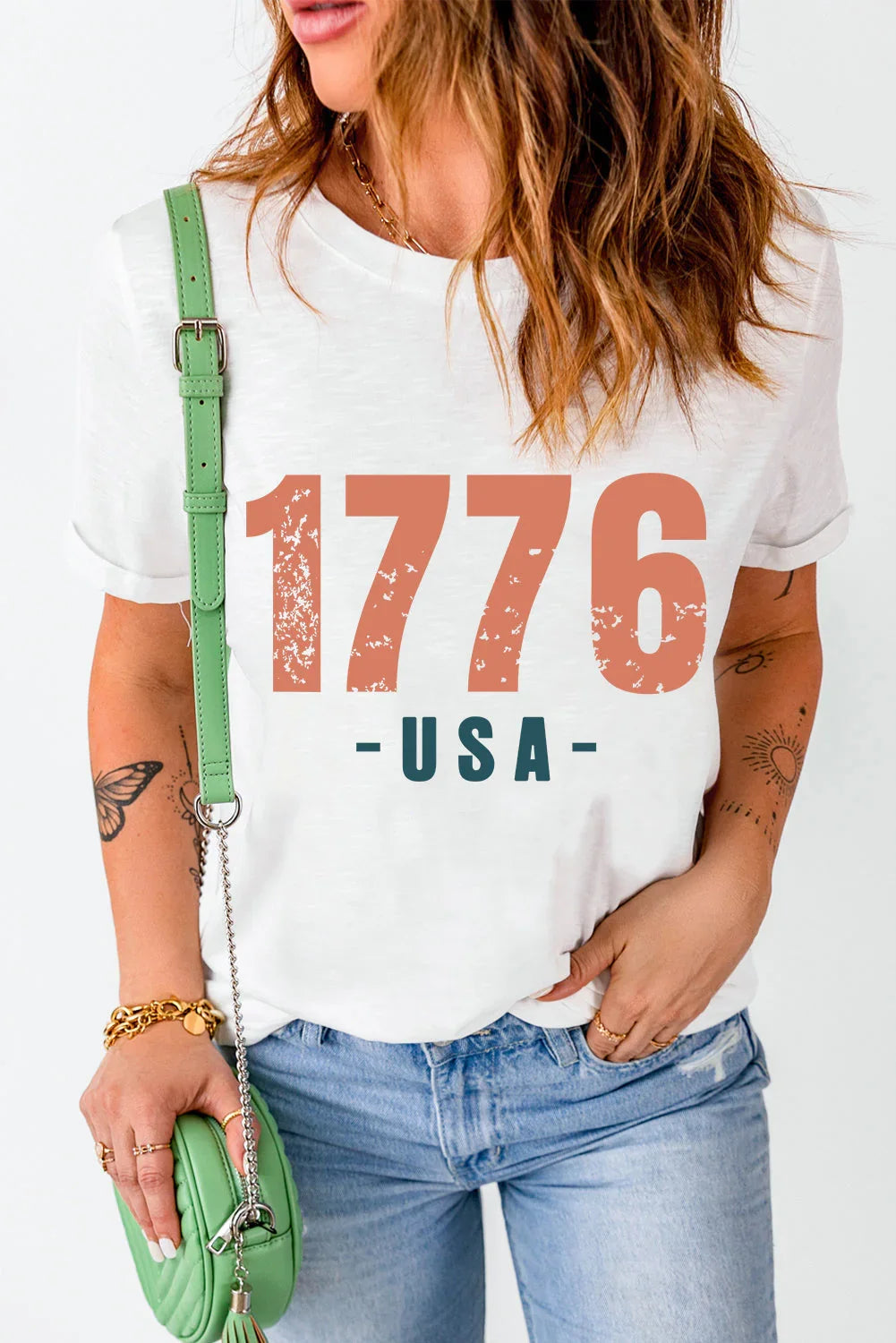 Woman wearing a white '1776 USA' graphic t-shirt with light blue jeans and a mint green crossbody bag