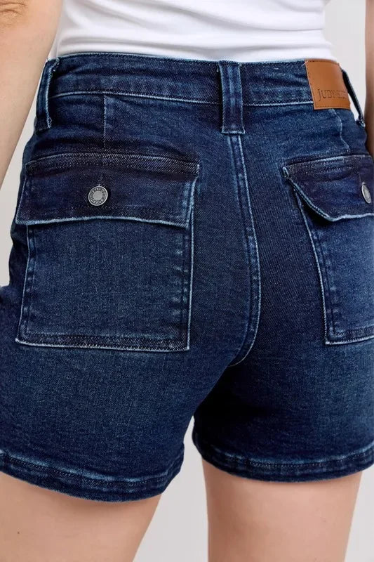 Back view of dark wash denim shorts with flap pockets, worn with a white top.