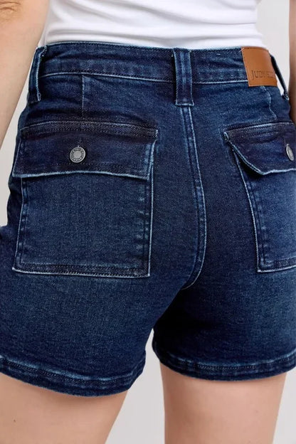 Back view of dark wash denim shorts with flap pockets, worn with a white top.