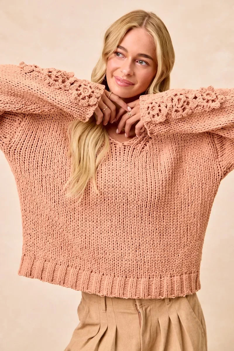 Woman wearing a blush pink chunky knit sweater with floral crochet sleeves and pleated beige pants.