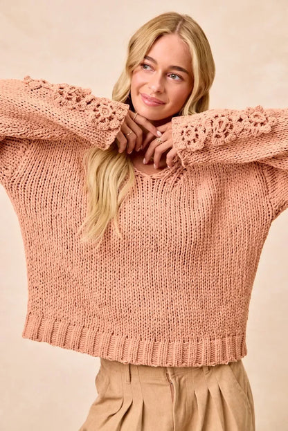 Woman wearing a blush pink chunky knit sweater with floral crochet sleeves and pleated beige pants.