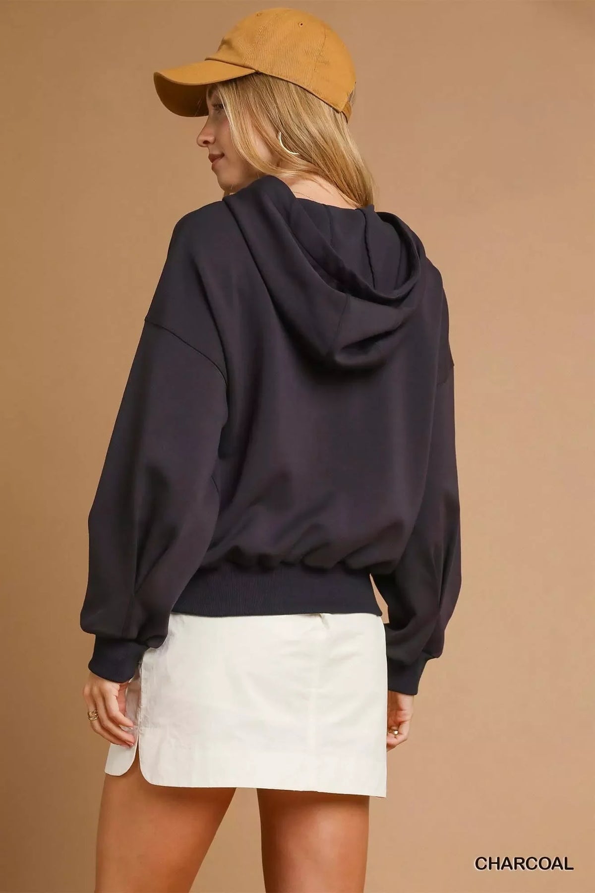 Woman in charcoal hooded sweatshirt, white mini skirt, and tan cap, back view