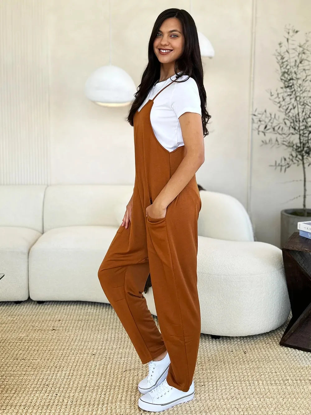Woman wearing rust brown relaxed jumpsuit over white t-shirt, white sneakers, modern living room setting.