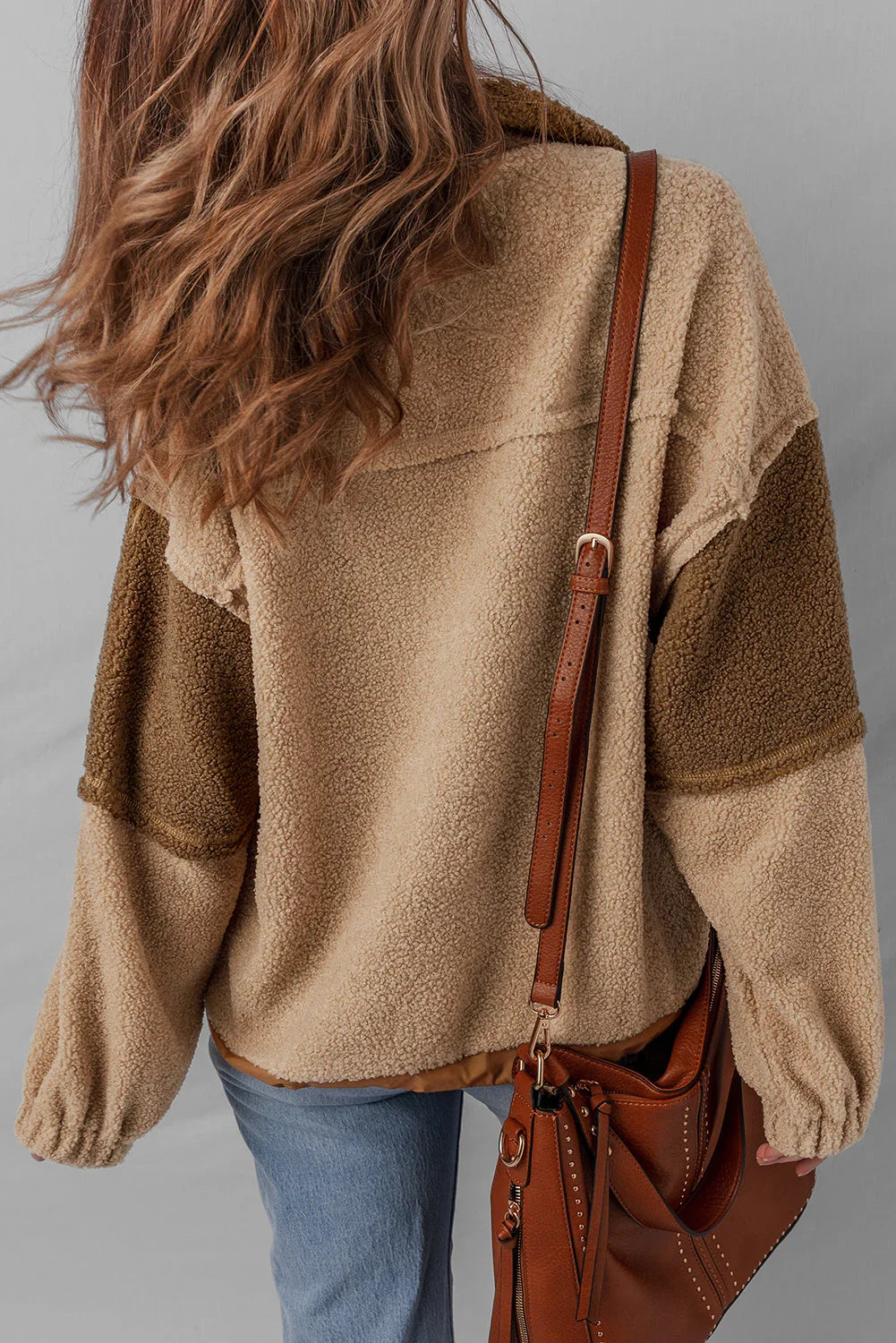 Woman in cozy tan sherpa jacket with color block sleeves, blue jeans, and brown leather bag