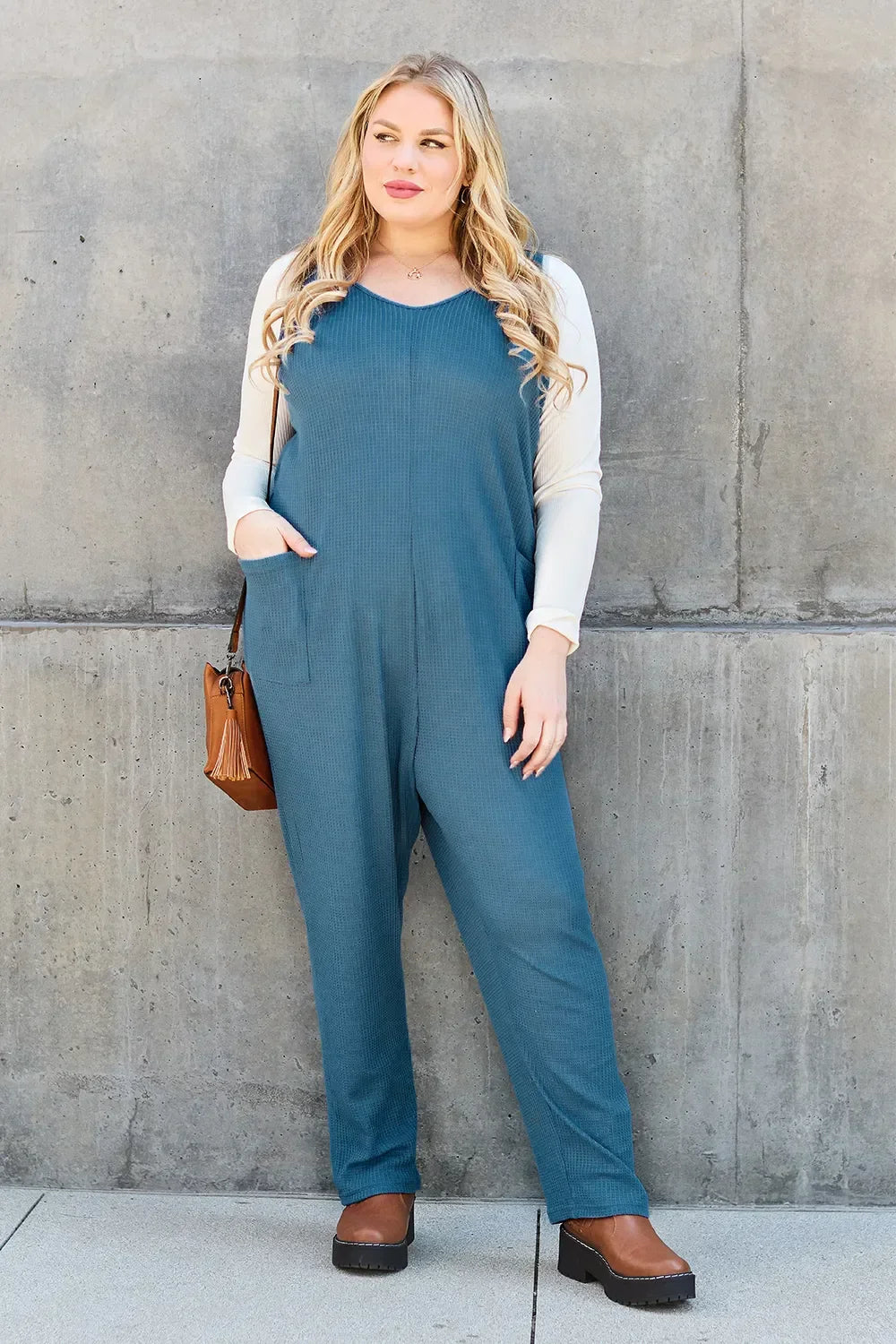 Woman in a blue relaxed-fit jumpsuit over a cream long sleeve top, brown shoes, and crossbody bag, standing against a concrete wall.