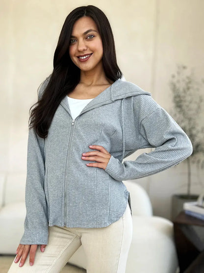 Woman modeling a light gray ribbed zip-up sweater with long sleeves and a relaxed fit indoors.