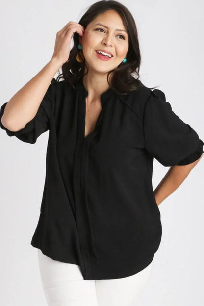 Woman wearing a loose black blouse with quarter sleeves and white pants, posing against a white background.