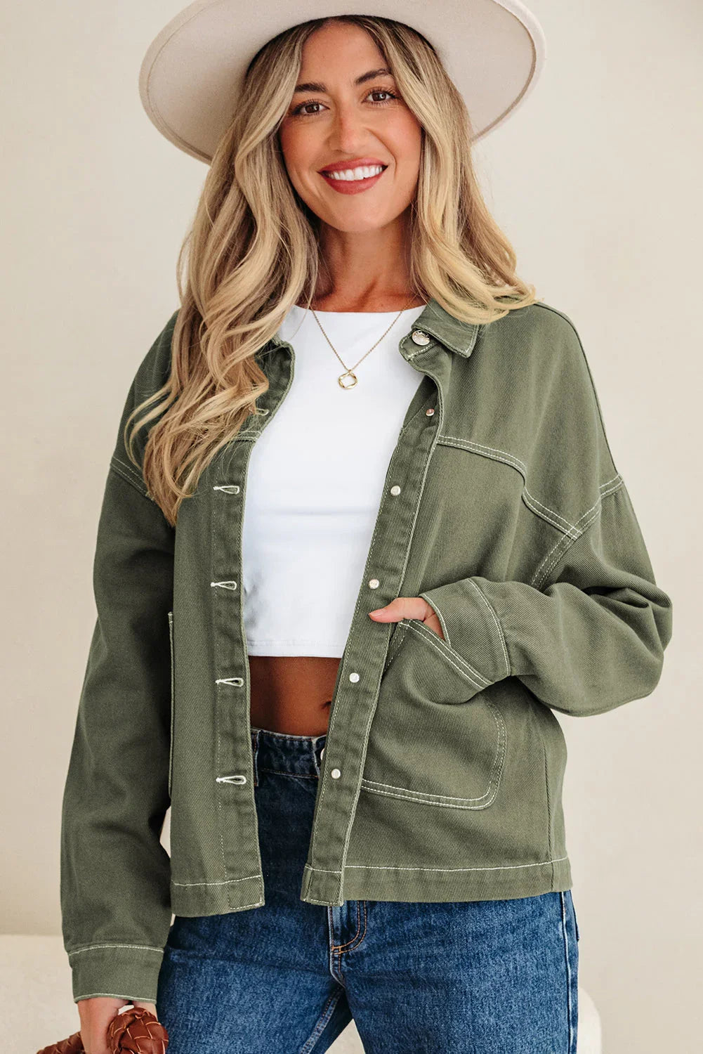 Woman in olive green jacket, white crop top, blue jeans, and wide-brim hat