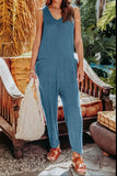 Woman wearing a relaxed blue sleeveless jumpsuit with pockets, holding a textured tote bag, standing outdoors near a wooden bench with tropical greenery.