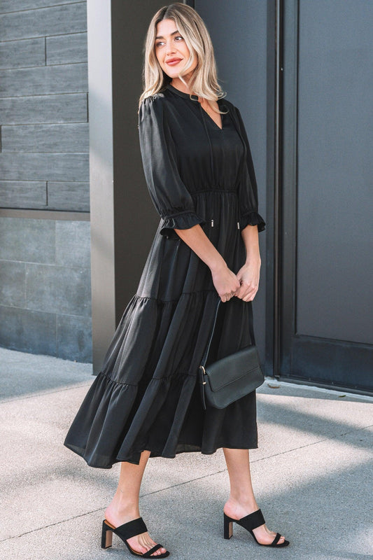 Woman in a black tiered midi dress with puff sleeves, holding a black clutch, wearing black heeled sandals