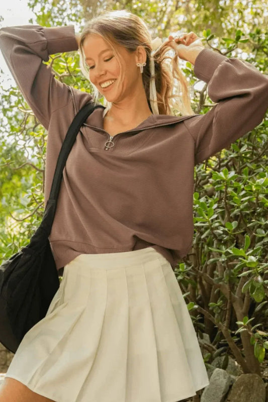 Woman outdoors in mauve zip pullover and white pleated skirt, smiling, with a casual bag