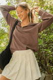Woman outdoors in mauve zip pullover and white pleated skirt, smiling, with a casual bag