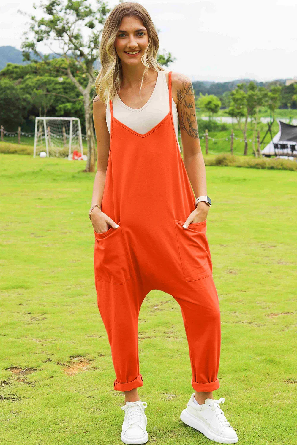 Woman wearing relaxed orange jumpsuit with side pockets, white tank top, and sneakers outdoors