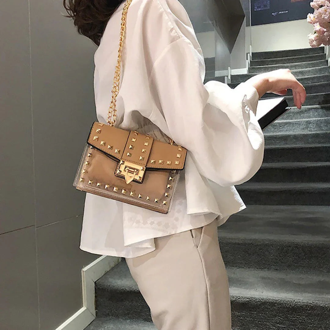 Tan clear shoulder bag with gold chain and stud detailing, worn by woman in white blouse