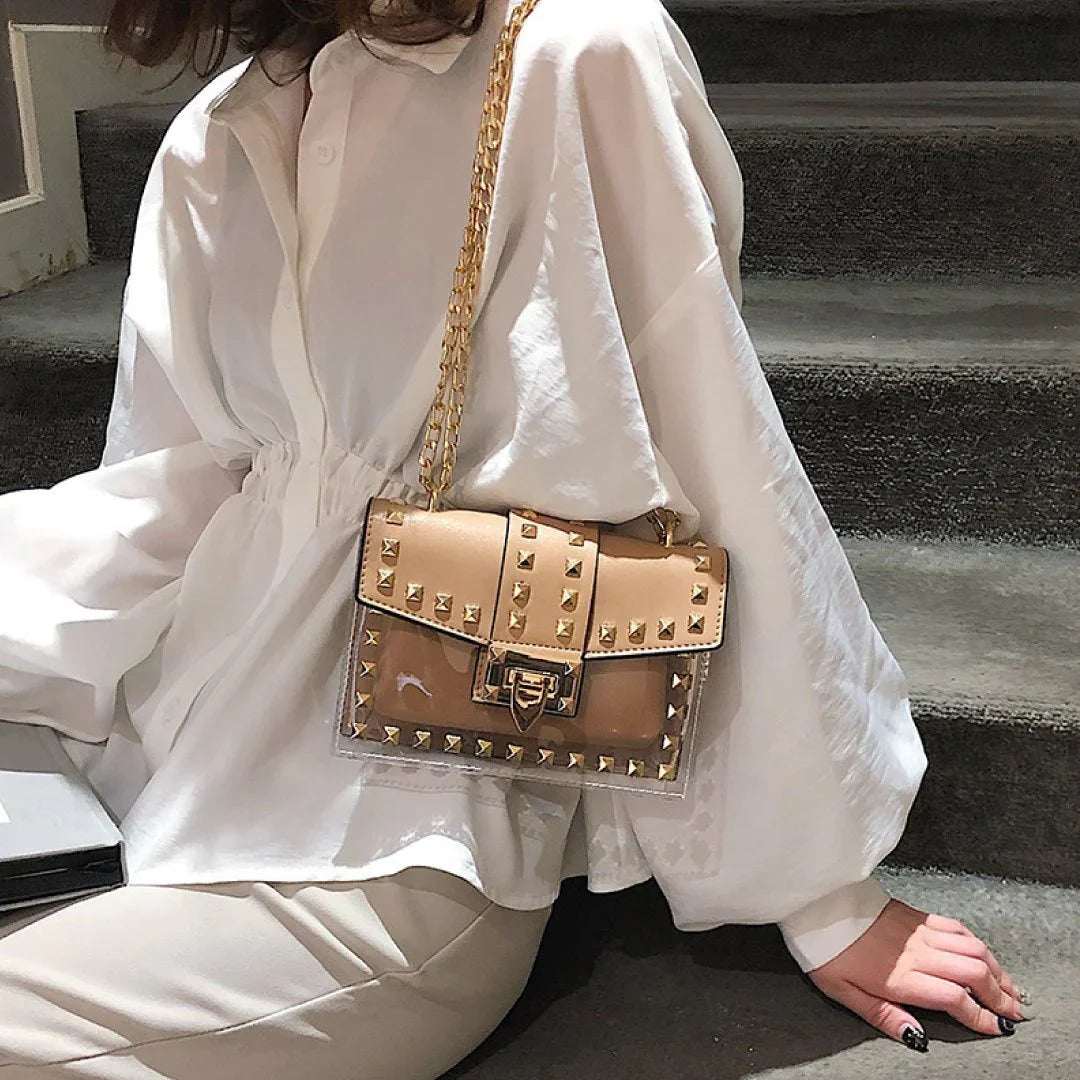 Tan clear shoulder bag with gold studs and chain strap, worn over a white blouse.