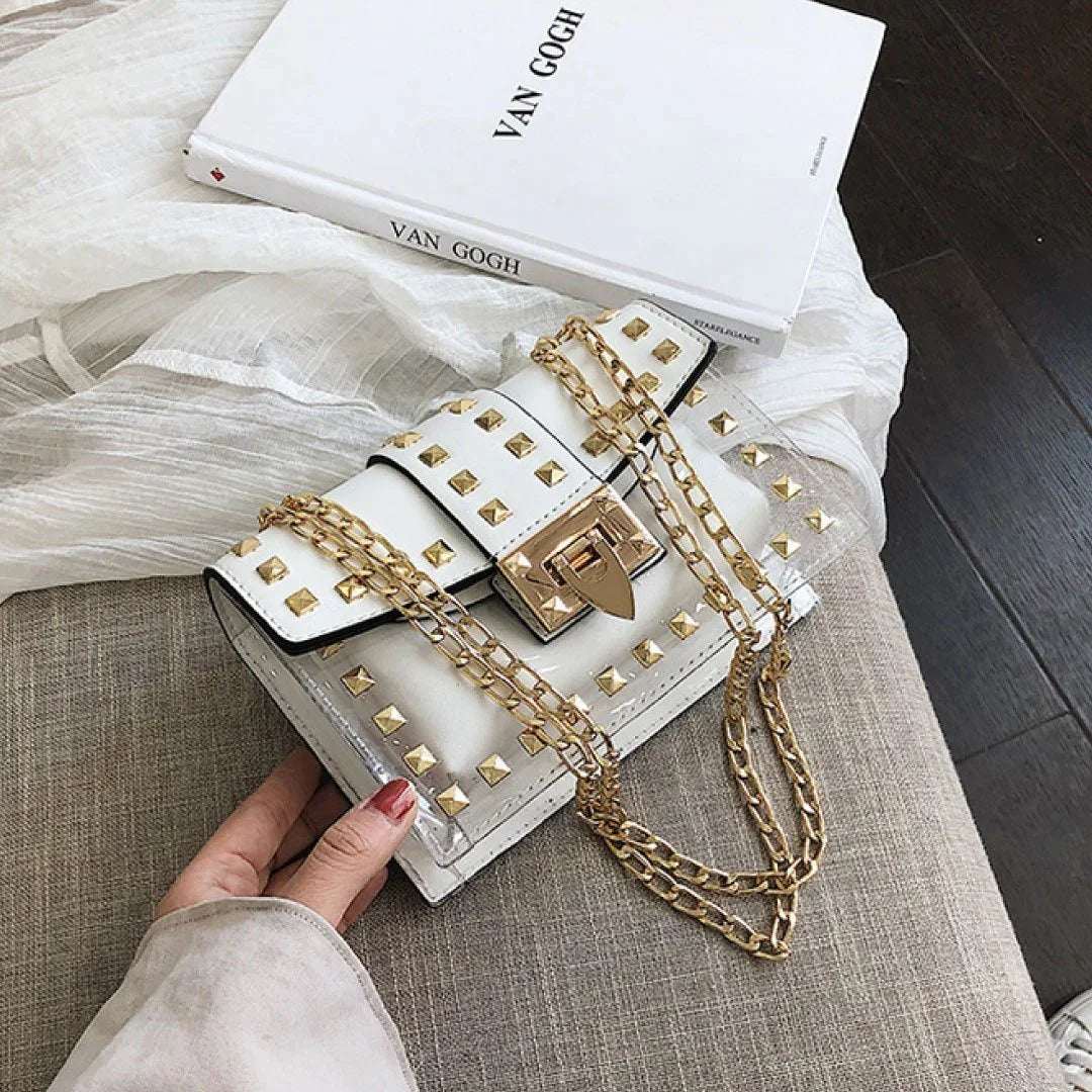 Clear and white studded shoulder bag with gold chain straps on a beige fabric surface