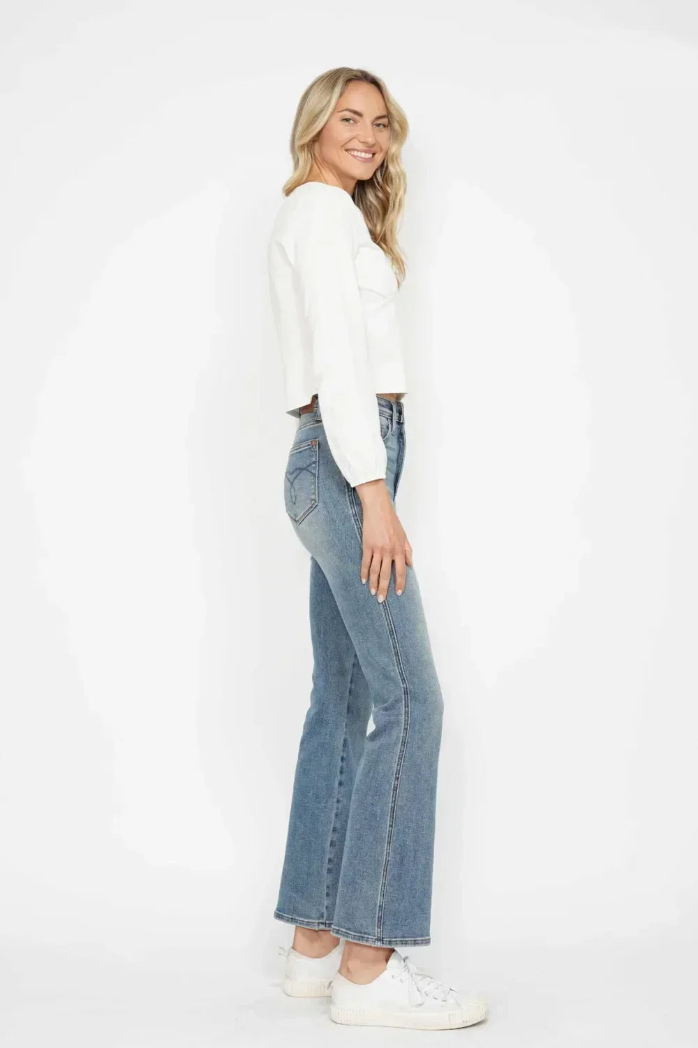 Woman in white long sleeve cropped top, high-waisted blue jeans, and white sneakers standing sideways against white background