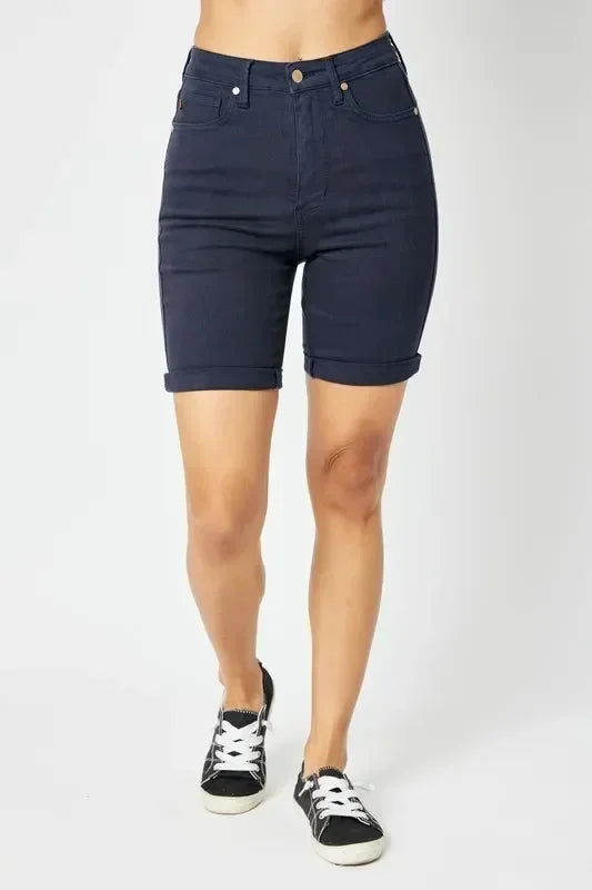 Woman wearing high-waisted dark denim Bermuda shorts with cuffed hems and black sneakers