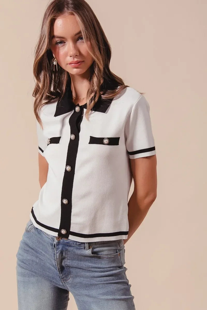 Woman wearing a chic short-sleeve white knit top with black trim and pearl buttons, paired with jeans.