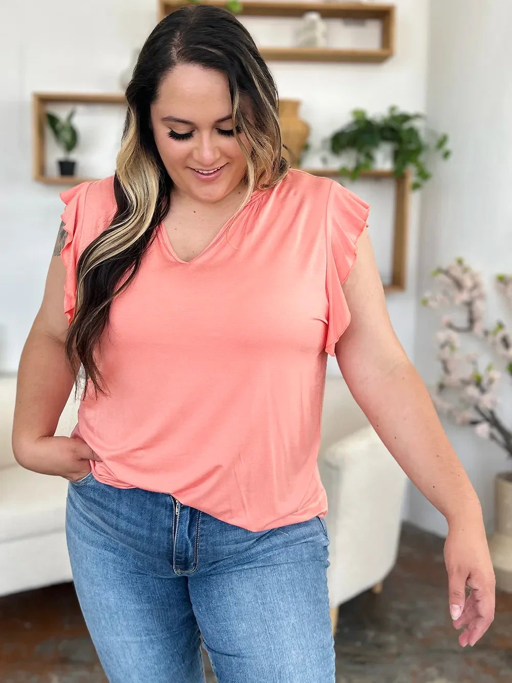 Woman wearing a coral flutter sleeve top and blue jeans in a modern living room setting