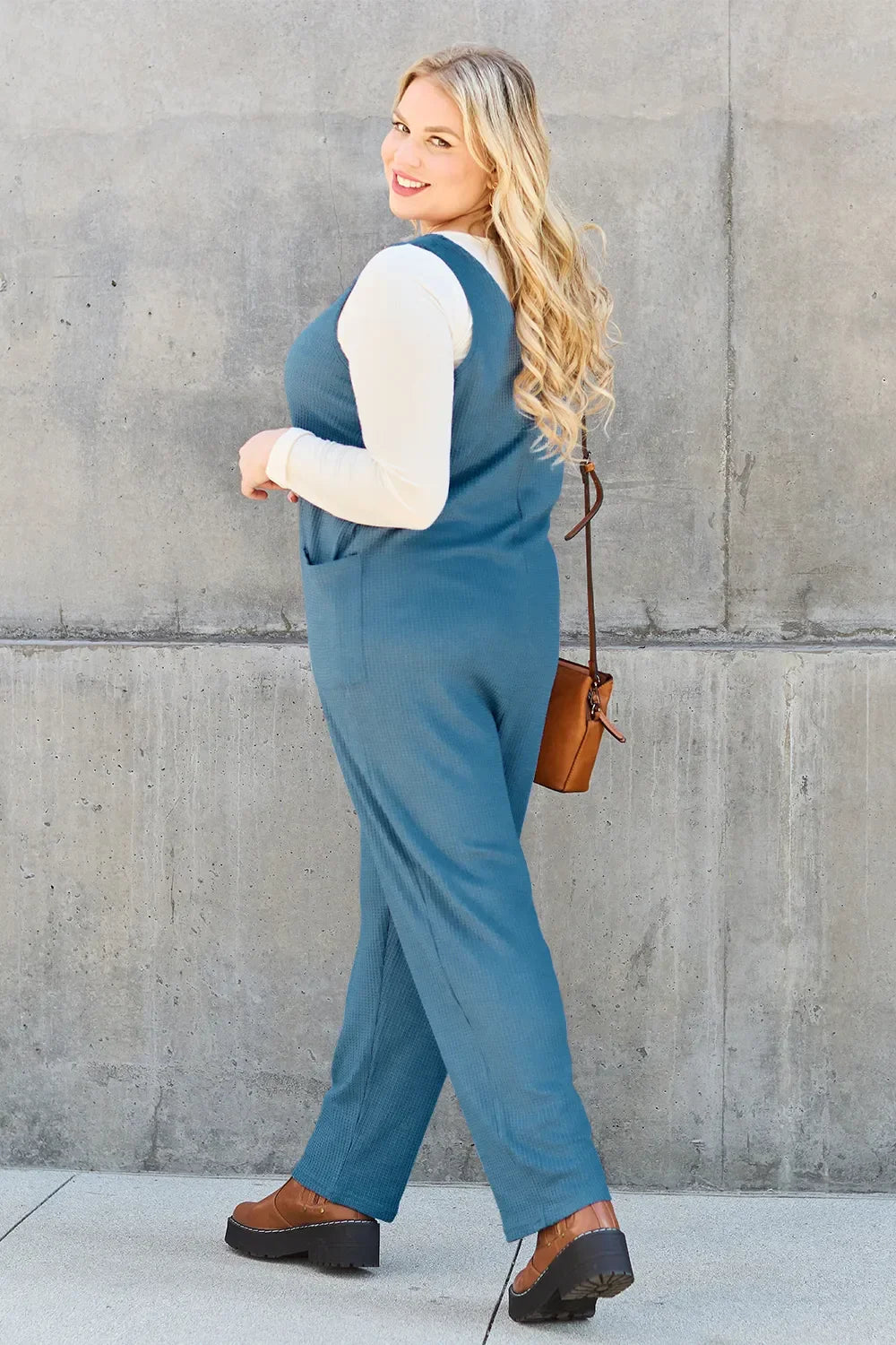 Woman in blue sleeveless jumpsuit, cream top, brown crossbody bag, and platform shoes