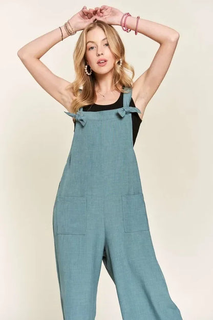 Woman wearing relaxed teal overalls with front pockets and tie straps over a black top