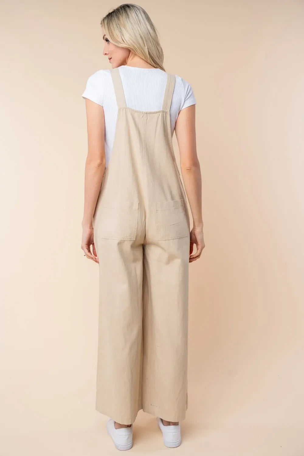 Woman in beige wide-leg overalls and white top, shown from the back, neutral background
