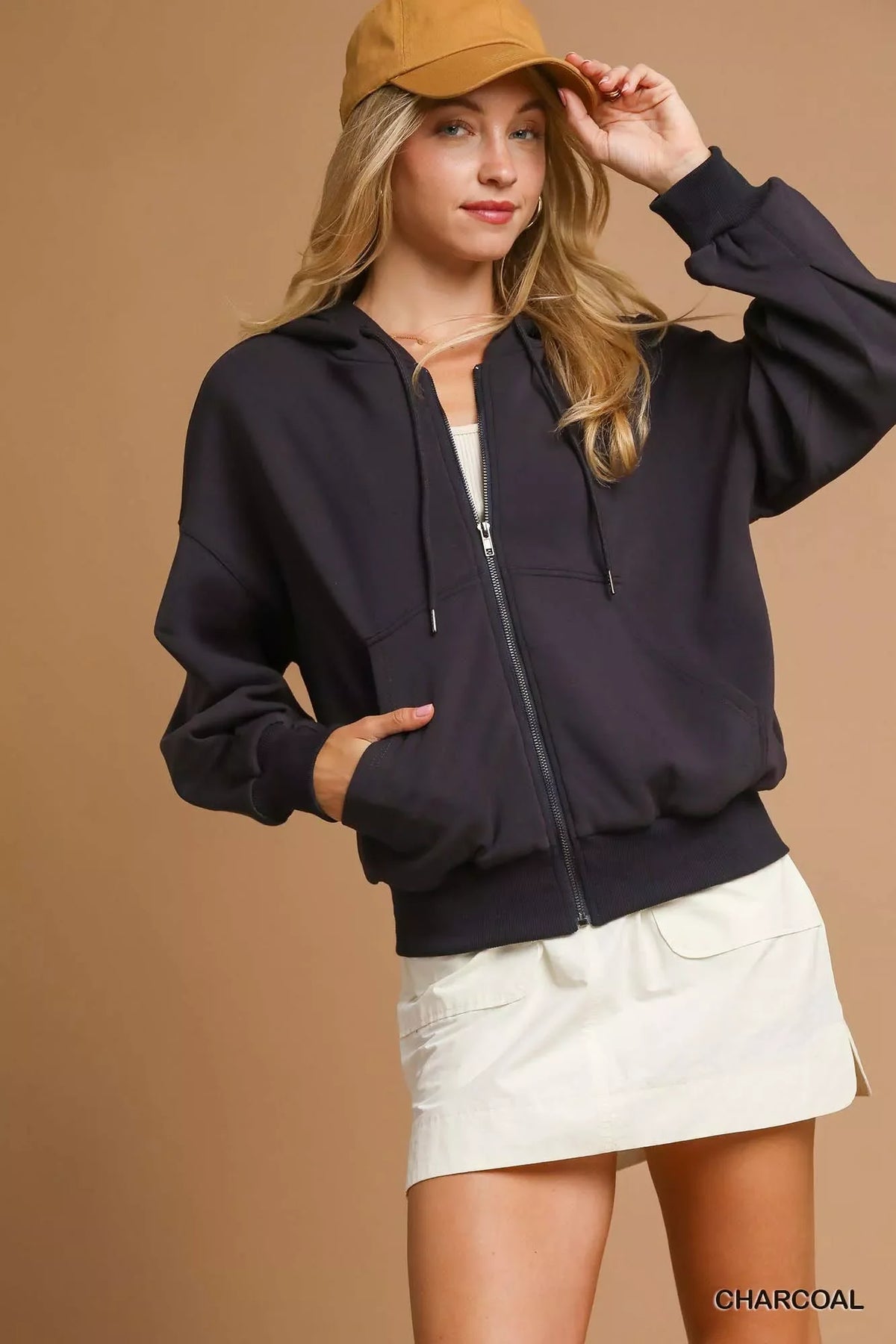 Woman wearing a charcoal zip-up hoodie, white skirt, and tan baseball cap against beige background