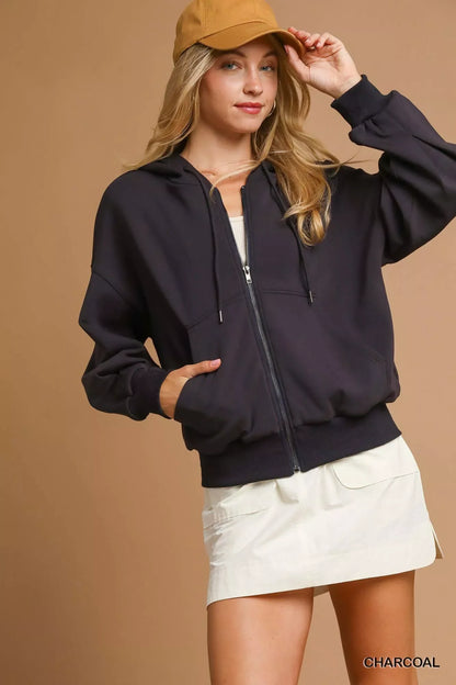 Woman wearing a charcoal zip-up hoodie, white skirt, and tan baseball cap against beige background