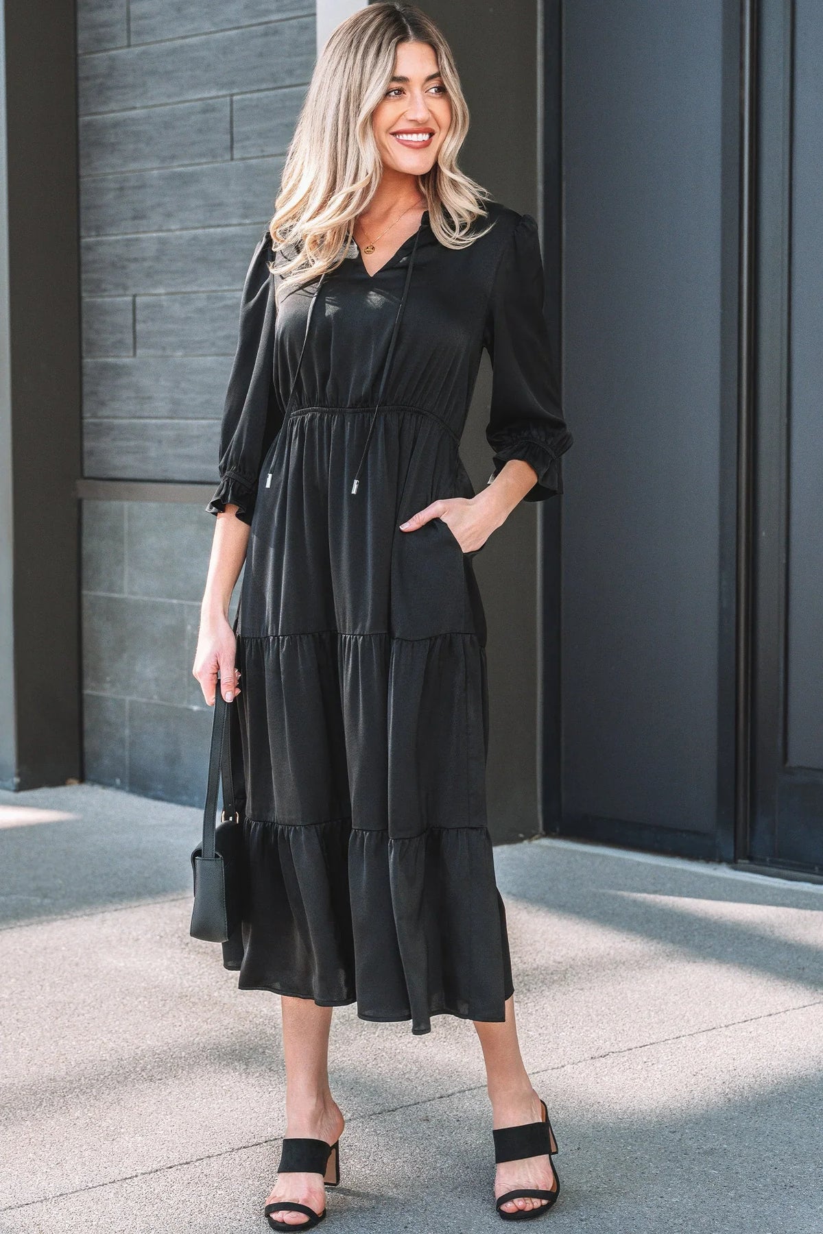 Woman wearing a black tiered midi dress with long sleeves, standing outdoors with a black handbag and sandals.