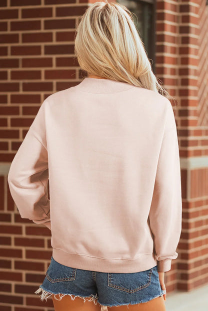Woman in a light pink sweatshirt and denim shorts standing outdoors against a brick wall
