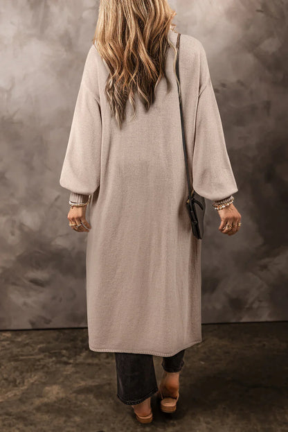 Woman wearing a long, taupe knit cardigan with relaxed sleeves and black handbag, seen from behind
