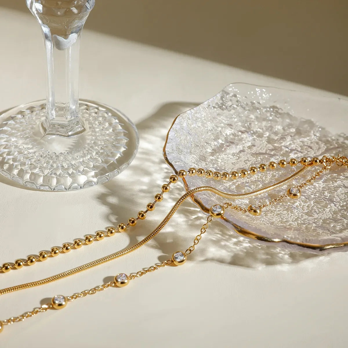 Gold layered necklaces with crystal accents on textured glass tray beside an elegant glass.