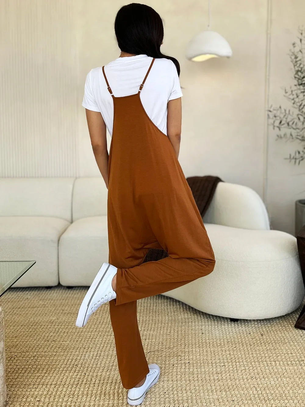 Woman in rust brown relaxed jumpsuit with white t-shirt and sneakers standing in modern living room