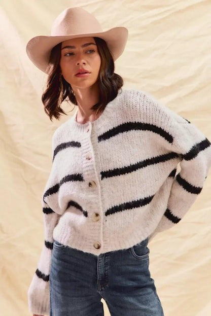 Woman in beige wool hat, striped button-up sweater, and blue jeans against neutral backdrop