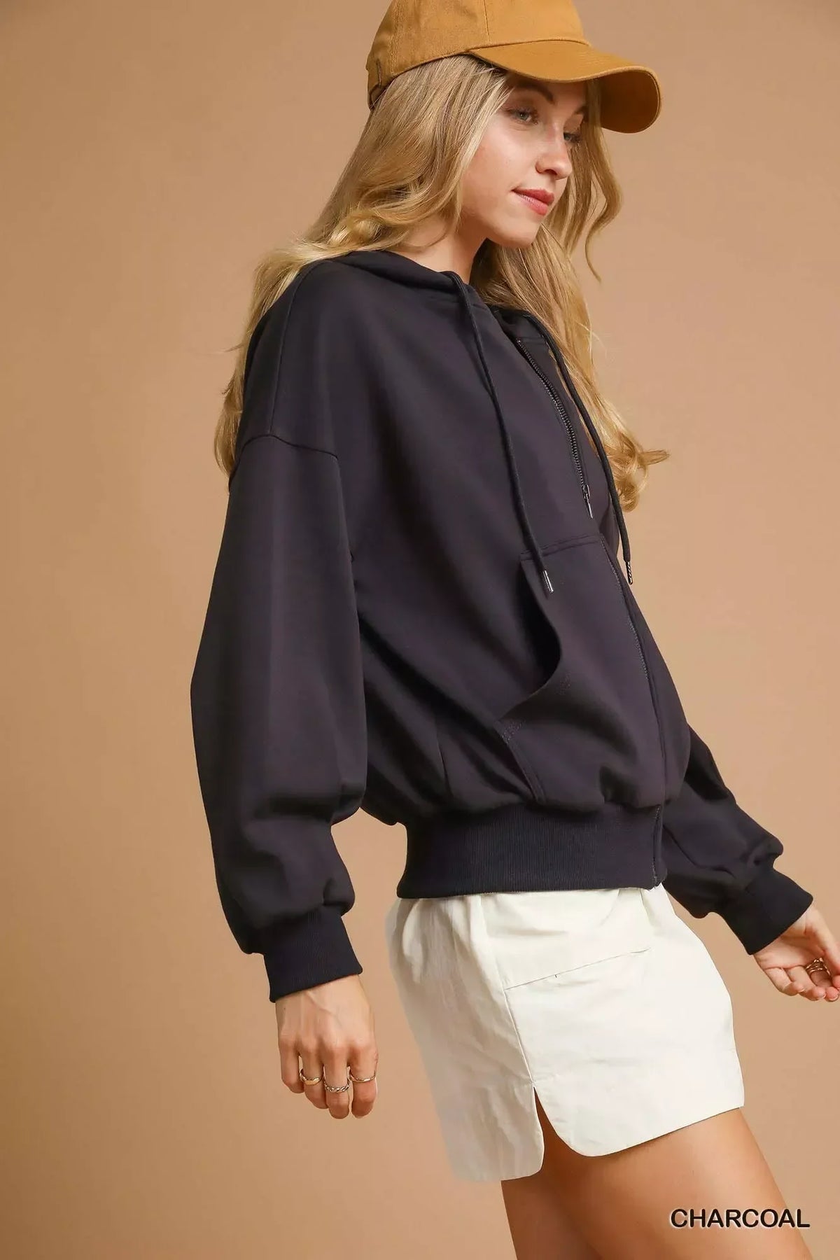 Woman wearing a charcoal hoodie, white shorts, and tan cap against beige background