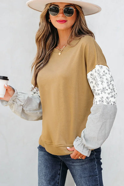 Woman in casual tan pullover with floral and striped sleeves, blue jeans, and a wide-brim hat