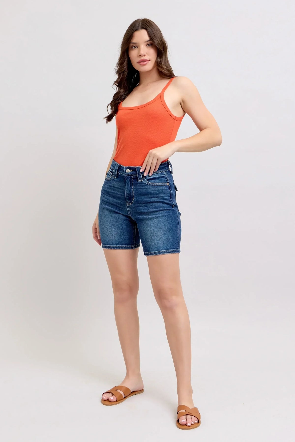 Woman wearing an orange tank top, denim shorts, and brown sandals on plain background