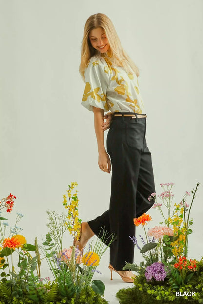 Woman in light floral blouse and black tailored pants, standing among colorful flowers