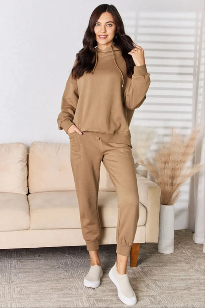 Woman wearing tan lounge set with hoodie and joggers, standing by beige sofa in modern living room