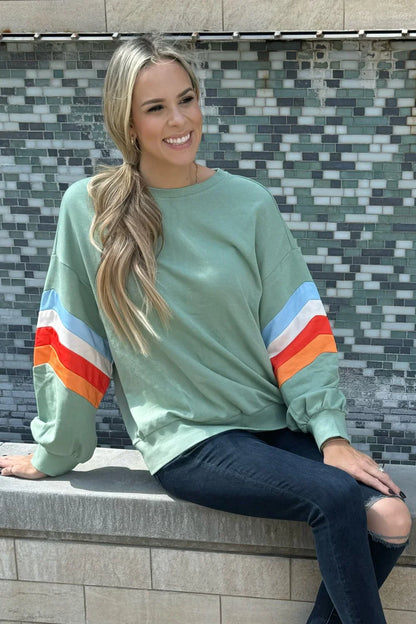 Woman in light green oversized sweatshirt with colorful striped sleeves, dark jeans, sitting outdoors