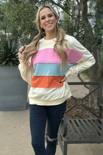 Woman wearing cream sweater with pink, blue, and orange colorblock stripes, outdoor setting